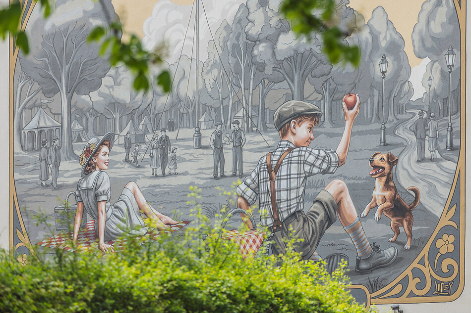 Mural przedstawia sielankową scenę w parku, gdzie na kocach piknikowych odpoczywają elegancko ubrana kobieta oraz chłopiec bawiący się z psem. W czarno-białym tle widoczne są spacerujące pary, drzewa oraz namioty festynowe, a dolną część kadru przesłaniają żywe, zielone krzewy.