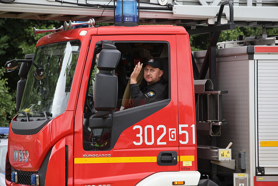 Zdjęcie przedstawia strażaka siedzącego w kabinie czerwonego wozu pożarniczego marki Iveco Eurocargo i wykonującego gest pozdrowienia ręką. Na drzwiach pojazdu wyraźnie widoczny jest numer operacyjny 302[G]51, a nad dachem kabiny zamontowano niebieską sygnaturę świetlną oraz fragment drabiny mechanicznej.