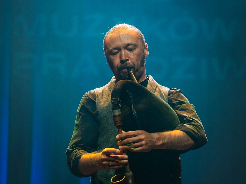 Grajek stoi na scenie i gra na instrumencie przypominającym dudę. Instrument ma duży, miękki worek trzymany pod pachą oraz drewnianą piszczałkę, w którą artysta dmucha. Osoba ma krótkie, jasne włosy i zarost na twarzy. Ubrana jest w ciemną koszulę oraz jasną kamizelkę. Oczy są zamknięte podczas gry. Tło jest jednolite i oświetlone w odcieniach niebieskiego, co podkreśla sylwetkę wykonawcy. Oświetlenie sceniczne skupia się na postaci, tworząc delikatne cienie na twarzy i instrumencie. W kadrze nie ma innych osób ani obiektów, a cała uwaga skierowana jest na muzyka i jego instrument.