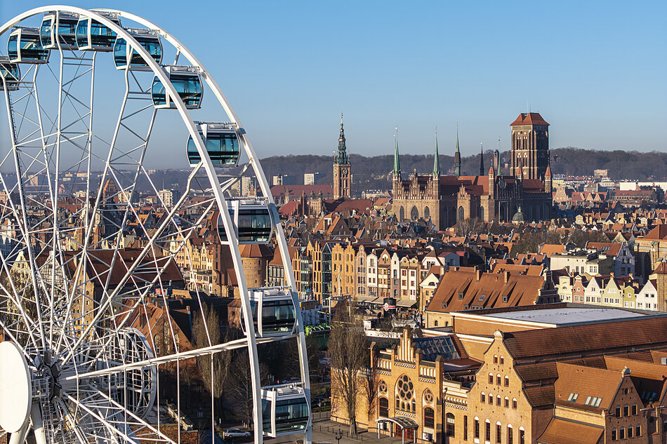 Zdjęcie przedstawia malowniczą panoramę Gdańska z widocznym na pierwszym planie kołem widokowym AmberSky i zabytkową architekturą Głównego Miasta. W tle górują charakterystyczne punkty orientacyjne, w tym monumentalna Bazylika Mariacka oraz wieża Ratusza Głównego Miasta.