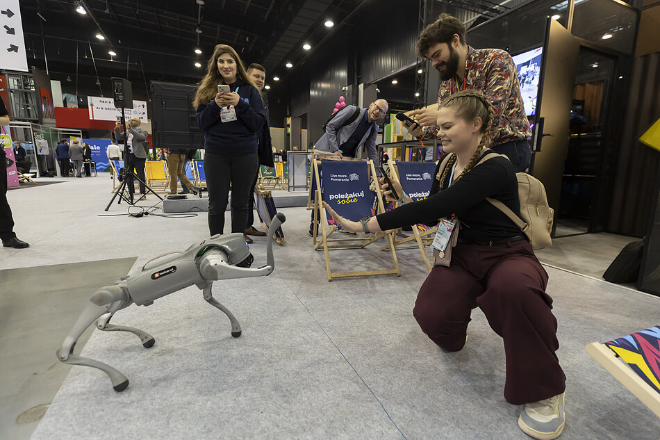 Zdjęcie przedstawia uśmiechniętą kobietę kucającą na targach i wyciągającą rękę w stronę zaawansowanego, czworonożnego robota przypominającego psa. W tle widać innych uczestników wydarzenia, którzy z zaciekawieniem obserwują i fotografują tę interakcję, siedząc lub stojąc przy leżakach z napisem „poleżakuj sobie”.