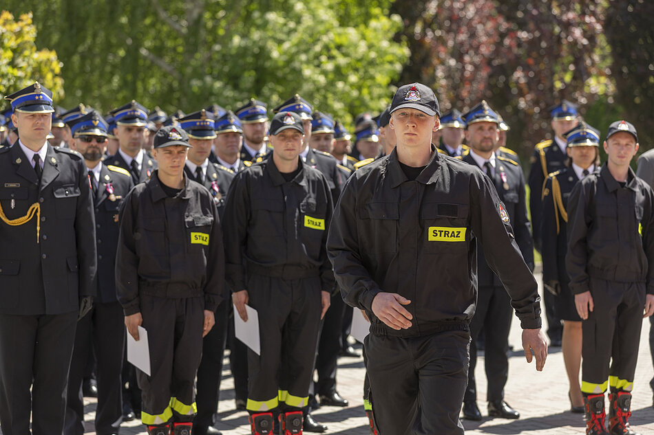 Zdjęcie przedstawia grupę umundurowanych strażaków stojących w zorganizowanym szyku podczas oficjalnej uroczystości na świeżym powietrzu. Na pierwszym planie widać idącego mężczyznę w czarnym ubraniu koszarowym z widocznym żółtym napisem „STRAŻ”.