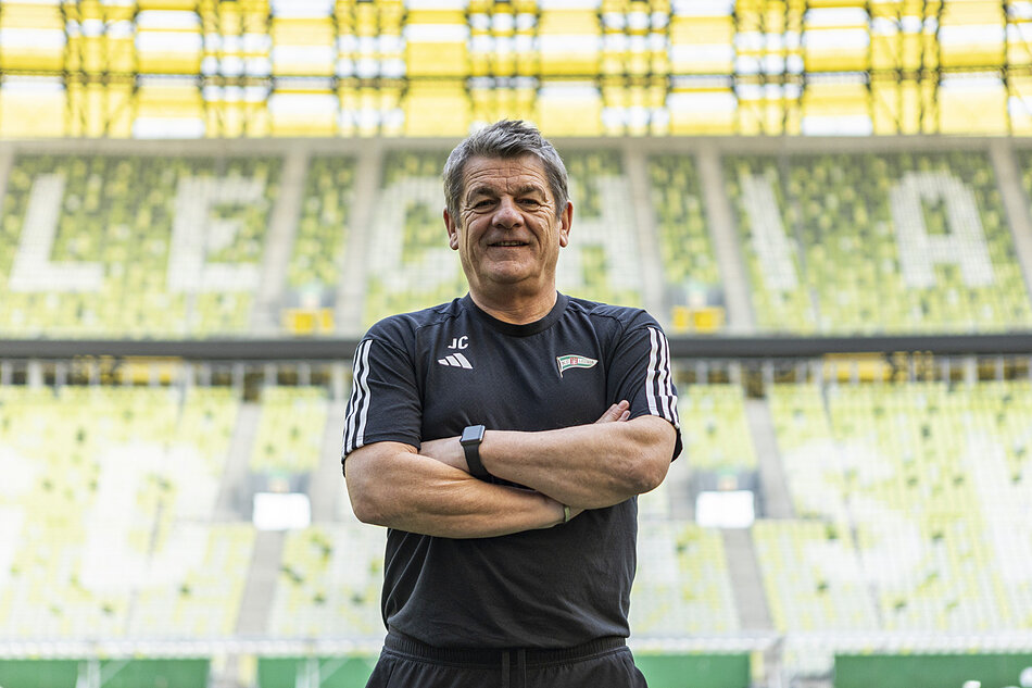 Mężczyzna lat około 60, w dresie, pozuje do zdjęcia na tle trybun stadionu
