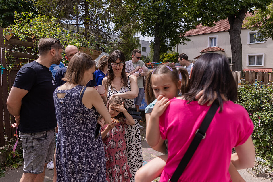 Otwarcie Klubu Sąsiedzkiego Przymorze Małe - grupa ludzi stoi na dworze i rozmawia ze sobą. Są kolorowo ubrani. W tle zielone rośliny.