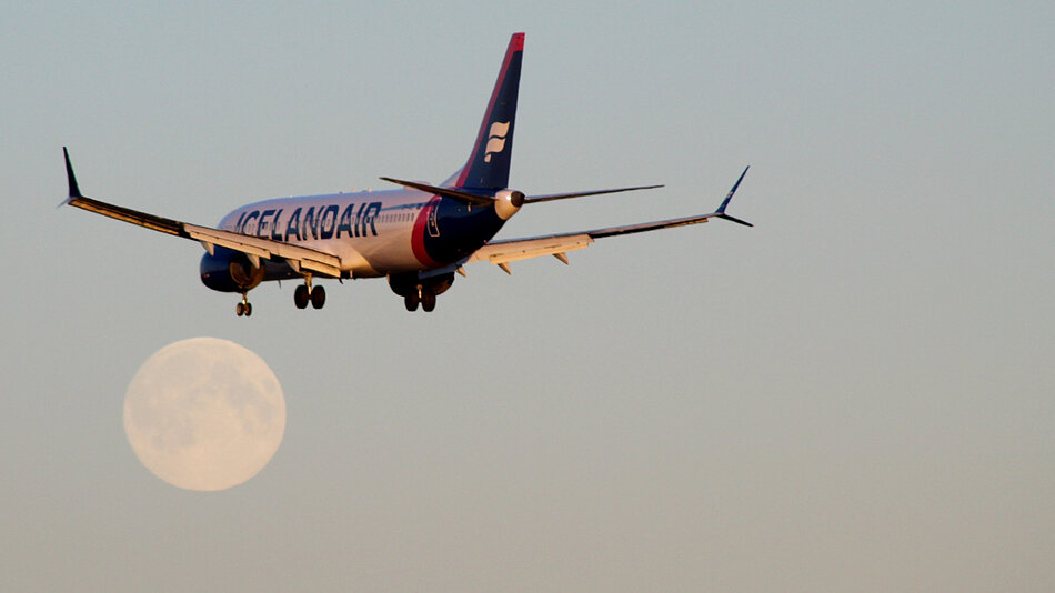 Zdjęcie przedstawia samolot linii Icelandair podchodzący do lądowania z wypuszczonym podwoziem. W tle, na jasnym niebie, widoczny jest wyraźny, pełny Księżyc.