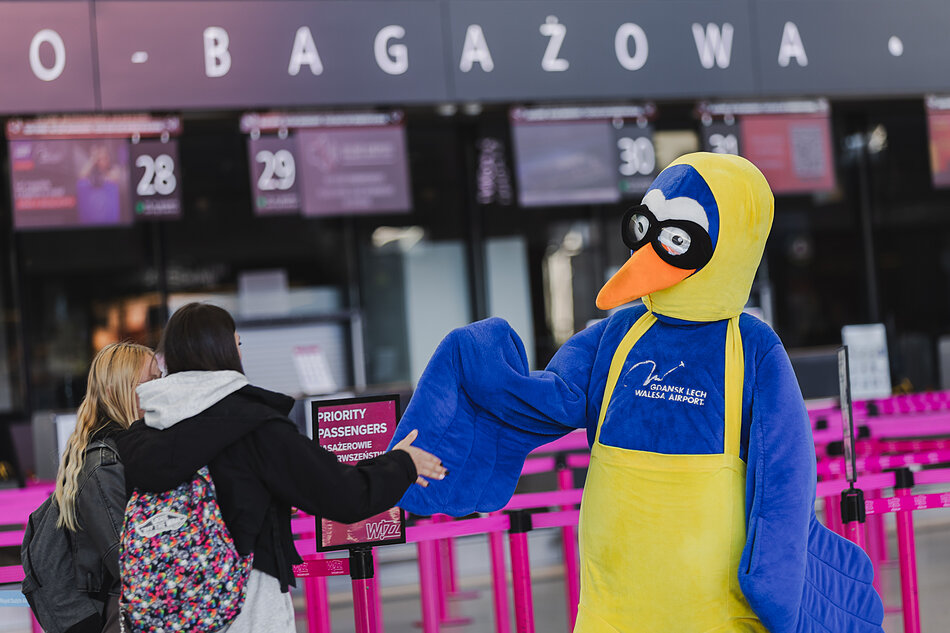Zdjęcie przedstawia maskotkę Portu Lotniczego Gdańsk im. Lecha Wałęsy witającą pasażerów w strefie odprawy bagażowej. Niebiesko-żółty ptak w okularach lotniczych podaje skrzydło kobiecie z plecakiem stojącej w kolejce.