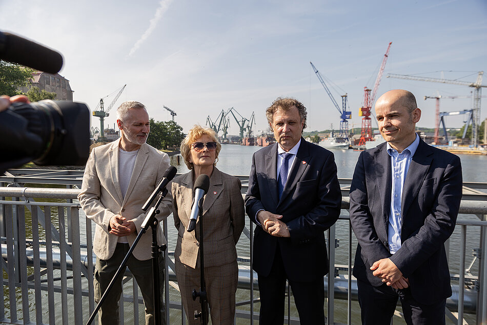 Jedna kobieta i trzech mężczyzn stoją przed mikrofonami na konferencji prasowej na moście. Za nimi rzeka, statki i dźwigi stoczniowe.