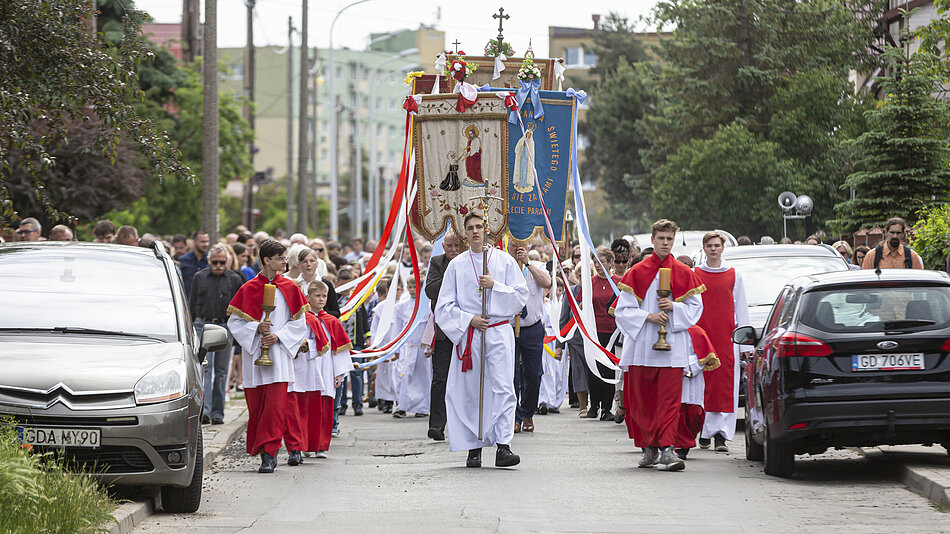 Duża grupa ludzi idzie ulicą niosąc religijne sztandary i krzyż, obok nich widać zaparkowane pojazdy