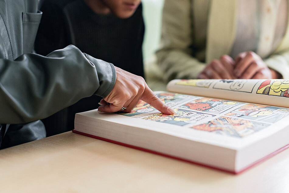 Zdjęcie przedstawia dłoń wskazującą konkretny kadr w otwartej, grubej księdze komiksowej leżącej na stole. Wokół książki widoczne są sylwetki osób, które wspólnie uczestniczą w czytaniu lub przeglądaniu ilustracji.