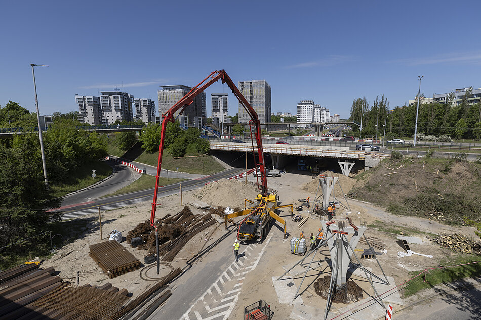 Zdjęcie przedstawia plac budowy infrastruktury drogowej w przestrzeni miejskiej, na którym widoczna jest duża pomarańczowa pompa do betonu z rozłożonym ramieniem. W tle wznoszą się nowoczesne wieżowce mieszkalne, a obok placu przebiega wiadukt z odbywającym się ruchem samochodowym.