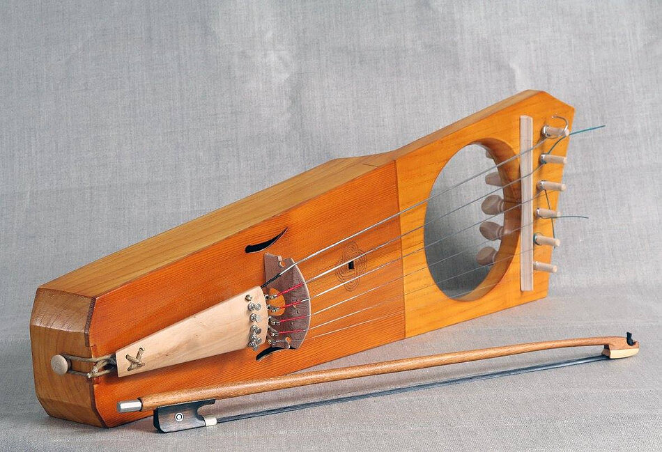 Zdjęcie przedstawia tradycyjny, smyczkowy instrument strunowy wykonany z jasnego drewna, charakteryzujący się podłużnym korpusem oraz dużym, okrągłym otworem rezonansowym. Obok instrumentu, na szarym tle, leży dopasowany smyczek, gotowy do gry.