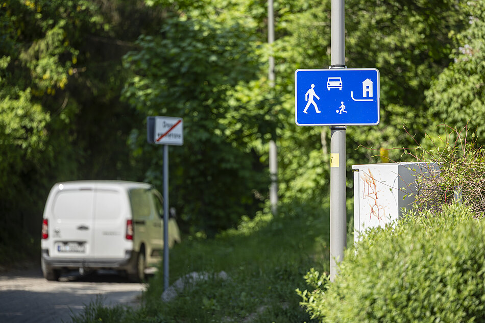 Zdjęcie przedstawia niebieski znak drogowy oznaczający strefę zamieszkania, umieszczony na słupku przy zielonym poboczu. W tle widać biały samochód dostawczy jadący wąską drogą otoczoną gęstą roślinnością.
