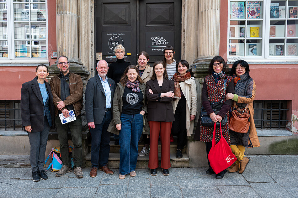 Grupa uśmiechniętych osób stoi przed wejściem do budynku z widocznymi napisami „Gdańsk Miasto Literatury” oraz „Tajne Komplety”. Uczestnicy spotkania są ubrani w codzienne, wiosenne stroje i pozują wspólnie na tle zabytkowych drzwi oraz okien wystawowych.