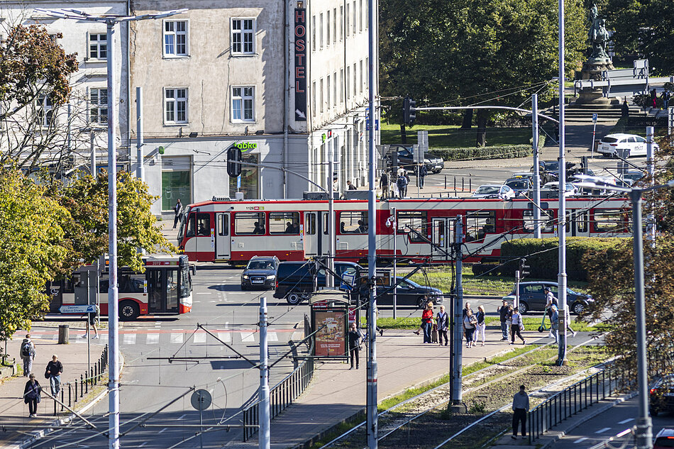 zdjęcie ulicy, widać jezdnie w obu kierunkach, jadące nimi samochody, jest też trakcja tramwajowa i chodniki, w tle widać przejeżdżający biało czerwony tramwaj