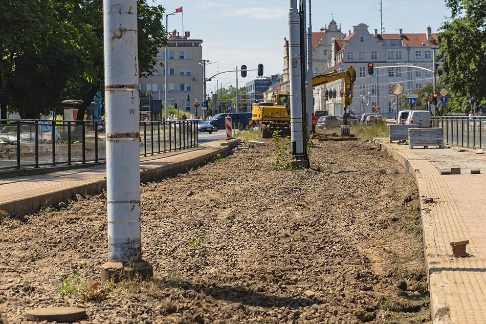 rozkopany teren gruntowy, widać słupy sieci trakcyjnej, w tle samochody i budynek urzędu po lewej, 