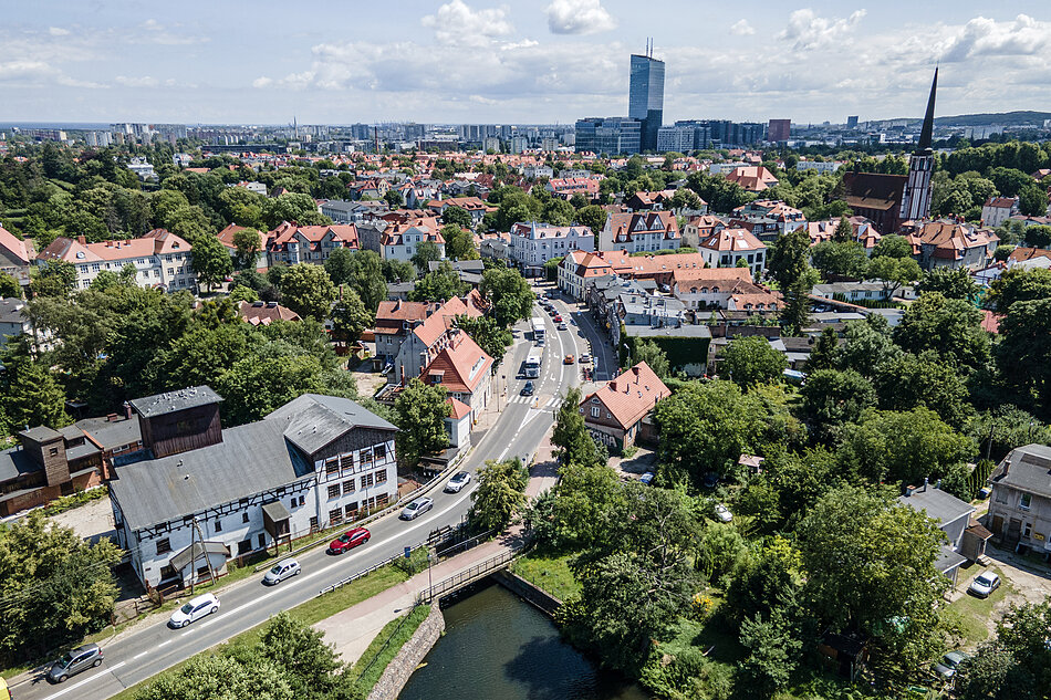 zdjęcie z drona, widać dzielnicę oliwę, czyli niewysokie kamienice z czerwonymi spadzistymi dachami, jest dużo zielonych drzew, w tle wysoki wieżowiec oszklony