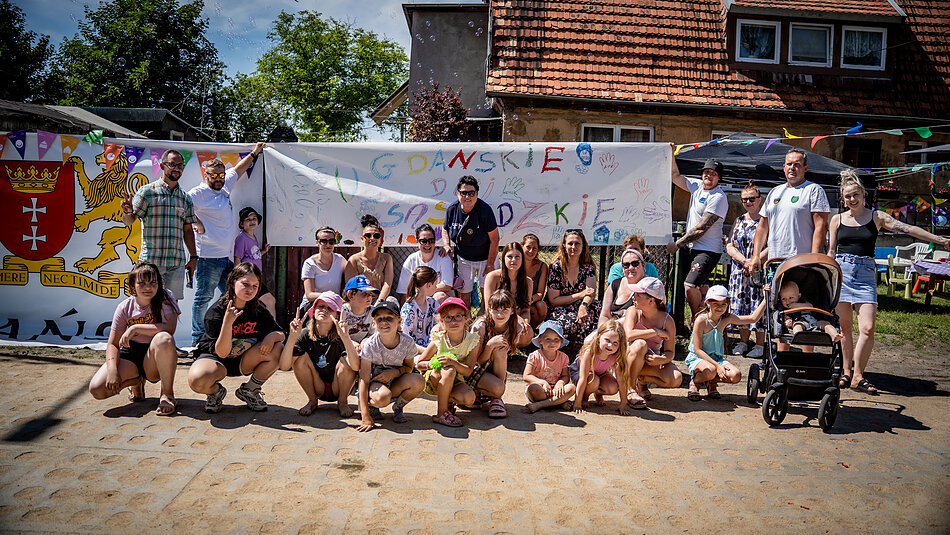 Zdjęcie przedstawia dużą grupę dorosłych i dzieci pozujących do wspólnej fotografii na świeżym powietrzu w słoneczny dzień. W tle widoczny jest dom jednorodzinny oraz rozciągnięty biały transparent z napisem „Gdańskie Dni Sąsiedzkie” i herbem Gdańska po lewej stronie.