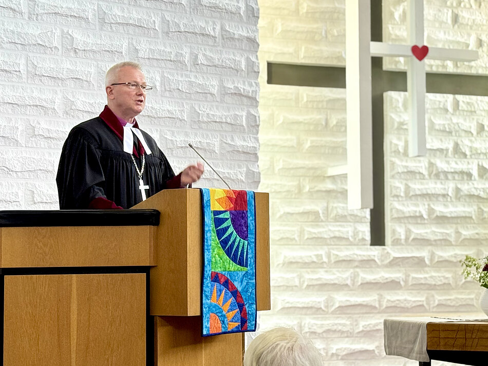 Zdjęcie przedstawia duchownego w okularach i liturgicznej todze z białym kołnierzem, który przemawia z drewnianej ambony ozdobionej kolorową tkaniną. Mężczyzna znajduje się na tle jasnej, strukturalnej ściany przypominającej kamień, obok której widoczny jest duży krzyż z zawieszonym małym serduszkiem.