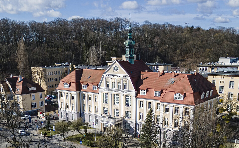 Zdjęcie przedstawia zabytkowy, wielopiętrowy gmach Gdańskiego Uniwersytetu Medycznego z czerwonym dachem i ozdobną wieżyczką. Budynek jest otoczony drzewami, a w tle widoczne są inne obiekty kampusu oraz zalesione wzgórze.