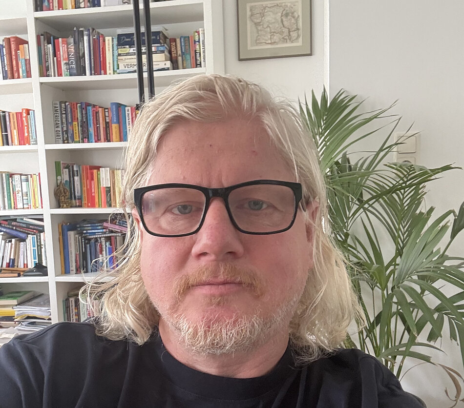 Portret osoby w czarnych okularach i ciemnej koszulce, sfotografowanej we wnętrzu mieszkania lub gabinetu. W tle widoczny jest biały regał wypełniony książkami, a po prawej stronie duża roślina doniczkowa. Na ścianie znajduje się oprawiona grafika lub mapa. Zdjęcie wykonano w naturalnym, jasnym świetle, a kadr obejmuje twarz i górną część sylwetki osoby siedzącej przed regałem.