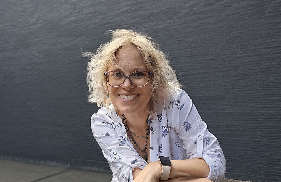 Portret osoby z krótkimi, jasnymi włosami i okrągłymi okularami, sfotografowanej na tle ciemnej, teksturowanej ściany. Osoba pochyla się lekko do przodu i opiera ręce przed sobą. Ubrana jest w jasną koszulę z motywem sów, nosi smartwatch oraz kilka naszyjników. Zdjęcie ma miękkie, naturalne oświetlenie, a tło jest jednolite i nie odciąga uwagi od głównego obiektu fotografii.