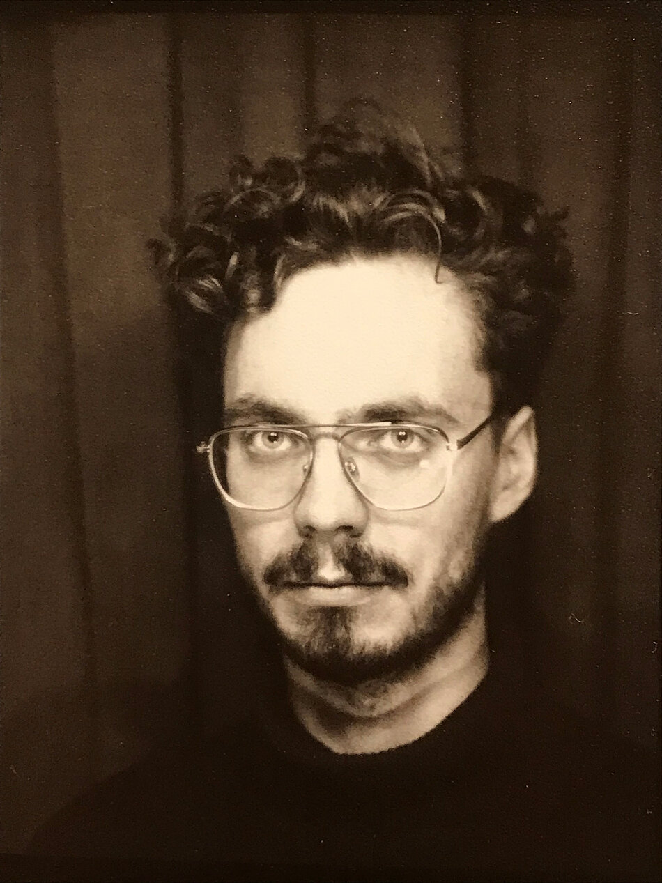 Czarno-biały portret przedstawiający osobę sfotografowaną od ramion w górę na tle ciemnej, materiałowej zasłony. Osoba ma krótkie, kręcone włosy, nosi duże okulary w cienkich oprawkach oraz zarost obejmujący wąsy i brodę. Ubrana jest w ciemny golf. Kadr jest zbliżeniem twarzy skierowanej w stronę aparatu, a fotografia utrzymana jest w klasycznej, kontrastowej stylistyce portretowej. Tło jest jednolite i nie zawiera dodatkowych elementów odwracających uwagę od postaci.