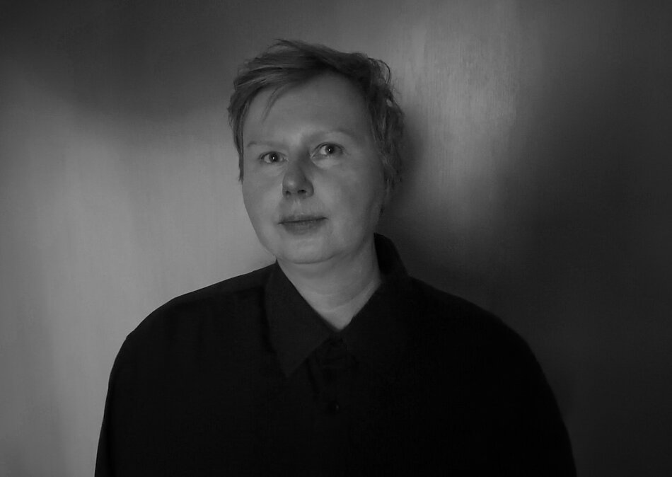 Czarno-biały portret osoby pokazanej od klatki piersiowej w górę. Osoba ma krótkie włosy i ubrana jest w ciemną koszulę z kołnierzykiem. Stoi na tle gładkiej, ciemnej ściany. Twarz jest oświetlona miękkim światłem, które tworzy delikatne przejścia tonalne i podkreśla rysy. Kadr jest prosty i oszczędny, skoncentrowany na postaci, bez dodatkowych elementów w otoczeniu. Zdjęcie utrzymane jest w monochromatycznej estetyce, z wyraźnym kontrastem między jasnymi partiami twarzy a ciemnym ubraniem i tłem.