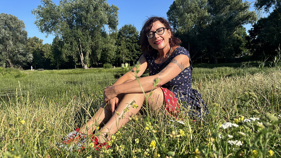 Opis alternatywny dla osób niewidzących: Osoba siedzi w wysokiej trawie na brzegu niewielkiego zbiornika wodnego w parku lub na terenach zielonych. Ma na sobie okulary, ciemną sukienkę w drobny wzór oraz czerwone obuwie. Ramiona opiera na zgiętych kolanach i zwraca się w stronę aparatu. Wokół rosną dzikie trawy i polne kwiaty, a w tle widać rozległą, zieloną przestrzeń oraz wysokie drzewa. Zdjęcie wykonano w słoneczny dzień, przy naturalnym świetle, które podkreśla intensywną zieleń roślin i błękit bezchmurnego nieba. Całość przedstawia spokojną scenę wypoczynku na łonie natury.