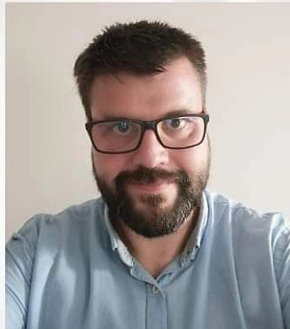 Portret osoby w okularach z ciemnymi oprawkami i zarostem, ubranej w jasnoniebieską koszulę. Zdjęcie wykonane na neutralnym, jasnym tle. Osoba znajduje się w centrum kadru i patrzy w stronę aparatu. Kadr obejmuje głowę oraz górną część tułowia.