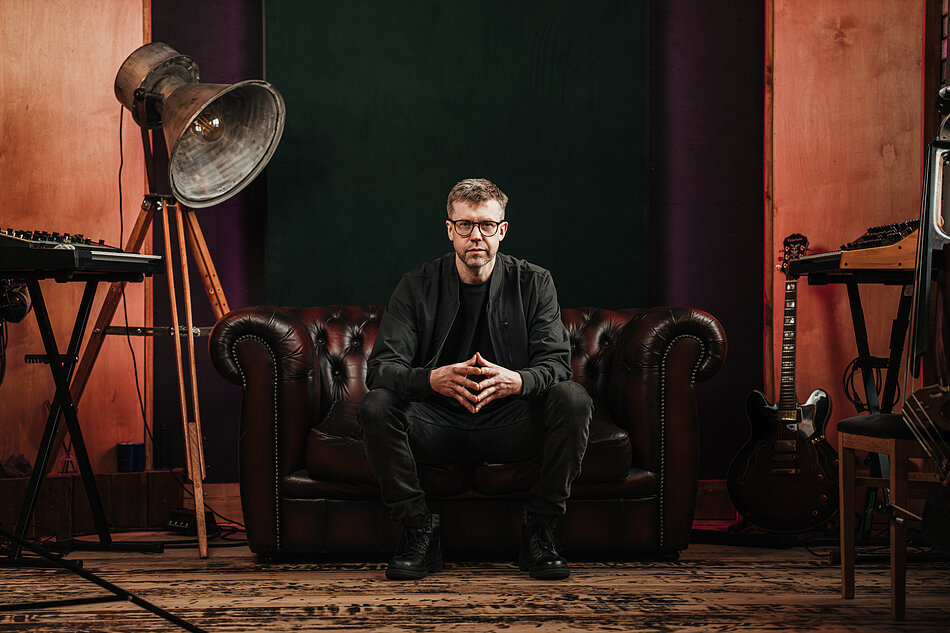 Osoba w ciemnym ubraniu i okularach siedzi na skórzanej, brązowej sofie typu chesterfield w studiu muzycznym. Po obu stronach widoczne są instrumenty muzyczne, w tym klawisze i gitara elektryczna. Obok sofy stoi duża lampa studyjna na drewnianym statywie. Wnętrze utrzymane jest w ciemnych, ciepłych barwach drewna, czerni i bordo, a tło tworzą panele akustyczne charakterystyczne dla przestrzeni nagraniowej.