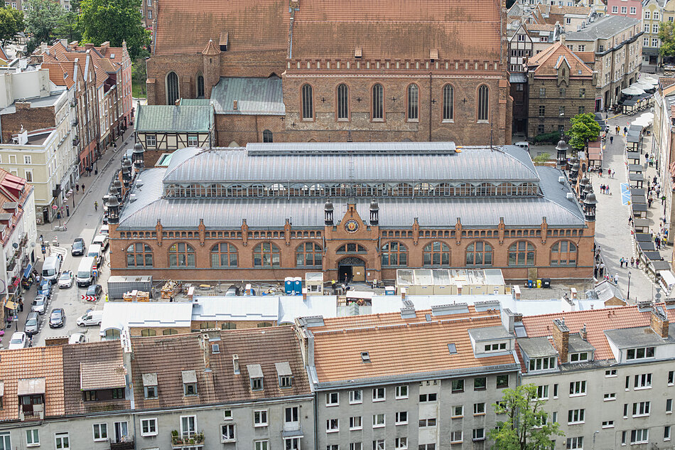  miasto z dużym budynkiem z czerwonej cegły w centrum. Budynek ma płaski dach i jest otoczony innymi budynkami z czerwonymi dachami płytowymi. W całym mieście rozsianych jest kilka sklepów i restauracji, a po prawej stronie obrazu widać ruchliwą ulicę z samochodami i ludźmi.