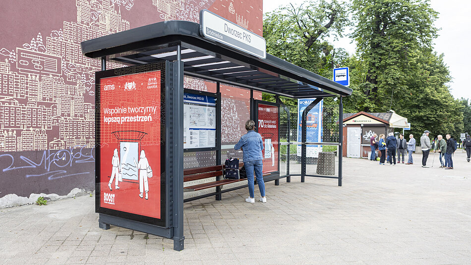  przystanek autobusowy w przestrzeni publicznej z czerwoną ścianą po lewej stronie. Ściana ma mural z pejzażem miasta z budynkami i drzewami. Po prawej stronie znajduje się niebieska tablica z napisanymi białymi literami słowami Przystanek Autobusowy poniżej tablicy z napisem Wiesbaden wiesnachtsmarkt co tłumaczy się na Witamy w mieście w języku angielskim. Jest również plakat z ilustracjami ludzi i tekstem na nim. Na pierwszym planie siedzący na ławce mężczyzna wydaje się czekać na swój autobus. Ma na sobie dżinsową kurtkę i jeansy i trzyma w ręku torbę. Za nim ludzie idą na chodniku, a w tle są drzewa i mały budynek.