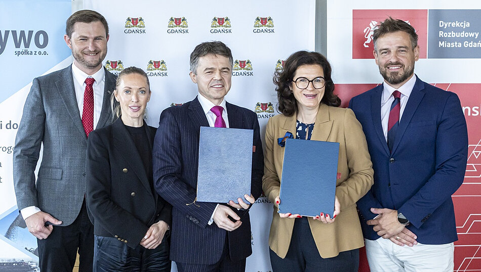Zdjęcie przedstawia grupę pięciu elegancko ubranych osób, które stoją na tle ścianki z logo miasta Gdańsk oraz Dyrekcji Rozbudowy Miasta Gdańska. Dwie osoby znajdujące się w centrum kadru trzymają w dłoniach oficjalne, ciemnoniebieskie teczki, co sugeruje moment podpisania ważnej umowy lub porozumienia.