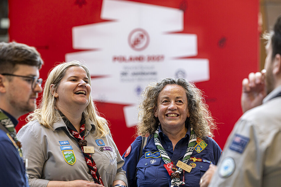 uśmiechnięci dorośli skauci w różnych mundurach, dwie kobiety i mężczyzna, w tle ścianka z logo World Scout Jamboree 2027 Poland