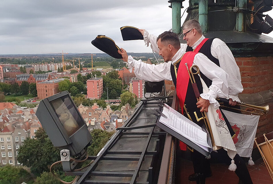 Zdjęcie przedstawia dwóch mężczyzn w historycznych strojach, którzy wychylają się z tarasu wieży i machają kapeluszami w stronę widocznego poniżej miasta. W tle rozciąga się panorama z zabytkowymi kamienicami, nowoczesną zabudową oraz zielonymi terenami pod pochmurnym niebem.