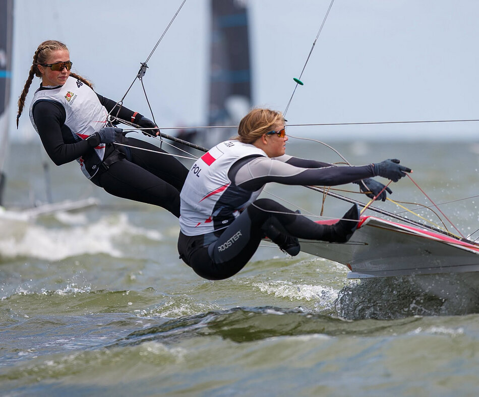 Zdjęcie przedstawia dwie sportsmenki w piankach i kamizelkach z napisem „POL”, które dynamicznie balastują na krawędzi żaglówki podczas rejsu po wzburzonej wodzie. Obie zawodniczki są mocno wychylone za burtę i skupione na precyzyjnym kontrolowaniu lin oraz stabilizacji płynącej łodzi.