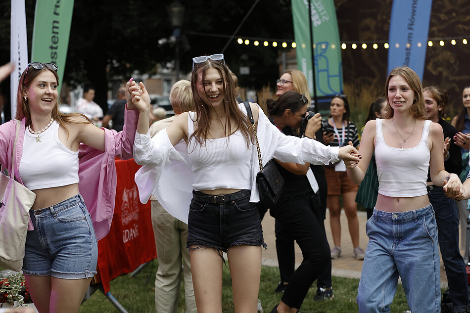 Trzy młode kobiety tańczące w parku, trzymające się za ręce i uśmiechające się do kamery, kobieta pośrodku nosi białą bluzę i dżinsowe szorty, a kobieta z lewej ma różową kurtkę na ramieniu, za nimi idą inni ludzie i stoją, można zobaczyć banery z tekstem, wydaje się, że kobiety są na festiwalu lub imprezie plenerowej.