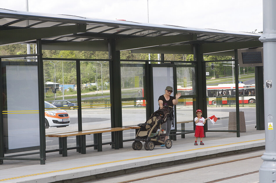 Przystanek autobusowy z kobietą pchającą wózek i dzieckiem stojącym na peronie; kobieta nosi czarną koszulkę, czapkę i okulary przeciwsłoneczne i macha do tłumu; trzyma czerwoną flagę w ręku, a dziecko stoi obok niej; po obu stronach kobiety stoją dwie ławki; w tle kilka autobusów zaparkowanych na dworcu autobusowym oraz drzewa i budynki widoczne przez okna.