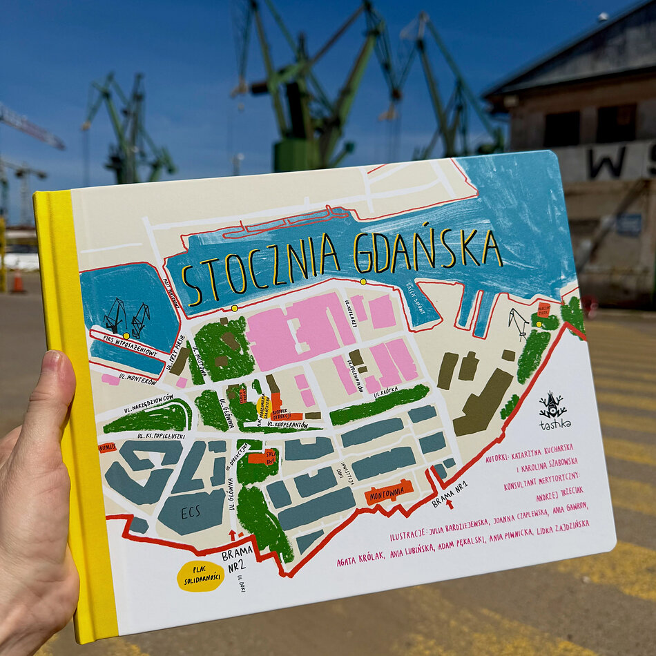 Zdjęcie przedstawia okładkę ilustrowanej książki dla dzieci zatytułowanej „Stocznia Gdańska”, wydanej przez wydawnictwo Tashka. Na pierwszym planie widoczna jest kolorowa mapa terenów stoczniowych z zaznaczonymi obiektami, takimi jak Europejskie Centrum Solidarności czy historyczne bramy.
