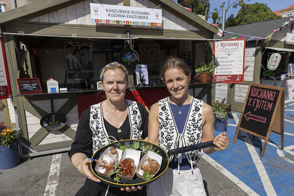 Dwie kobiety stojące przed stoiskiem spożywczym. Stoisko wykonane jest z drewna i ma napis Kaszubek Rückmann co tłumaczy się na Restauracja Rachael Raya. Kobieta po lewej nosi czarno-biały wzorzec i opaska głowy, a w rękach trzyma duży talerz z jedzeniem. Uśmiecha się i patrzy na aparat. Po prawej stronie jest młoda kobieta o blond włosach, również uśmiechnięta. W tle możemy zobaczyć inne stoiska spożywcze i budynki.