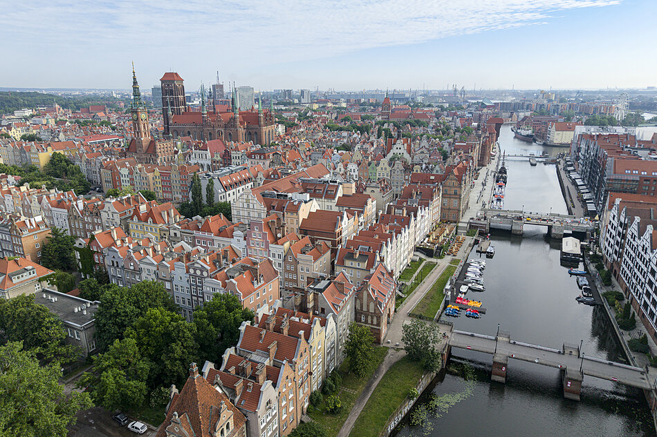 Miasto z rzeką biegnącą przez niego; miasto jest gęsto zaludnione czerwonymi domami i budynkami, a przez rzekę przechodzi kilka mostów; po prawej stronie most z kilkoma łodziami i jachtami zacumowanymi na wodzie; w tle widoczna jest panoramę miasta, z wysokimi budynami i wieżą zegarową w oddali; niebo jest niebieskie, i pogoda wydaje się słoneczna i przejrzysta.