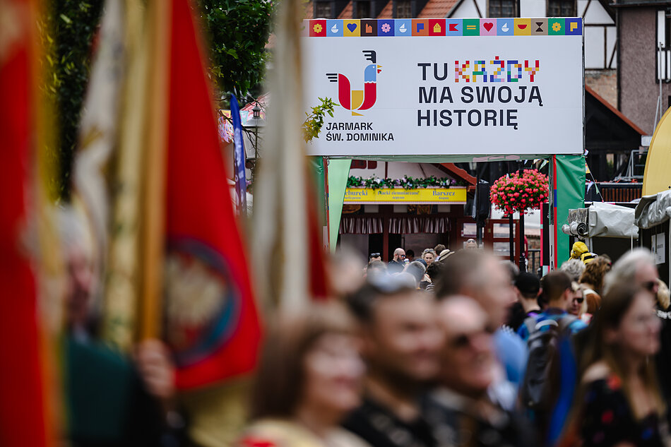 Zdjęcie przedstawia tłum ludzi zgromadzonych na tradycyjnym Jarmarku św. Dominika, nad którym góruje biały transparent z hasłem „Tu każdy ma swoją historię”. Na pierwszym planie widoczne są rozmyte, czerwono-białe polskie flagi, które nadają ujęciu uroczysty charakter.