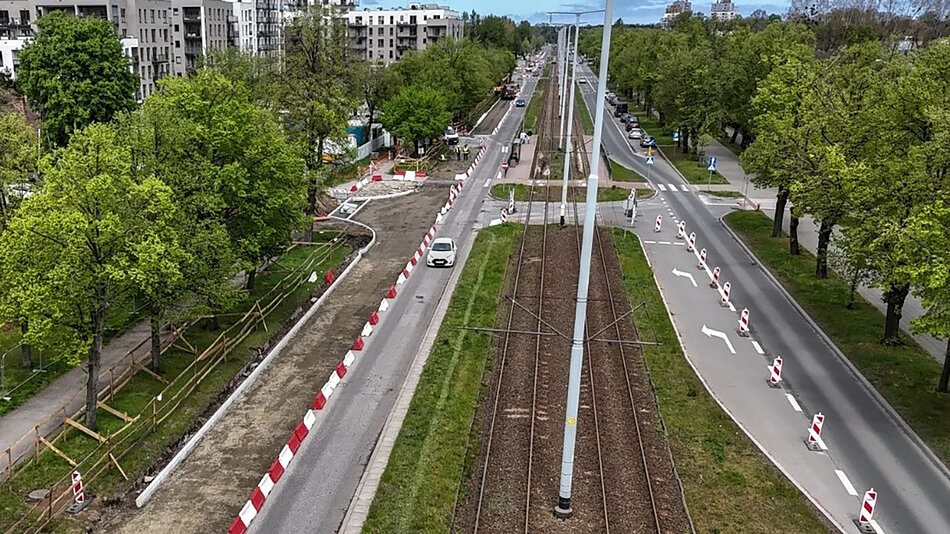 Zdjęcie z lotu ptaka na tory tramwajowe i jezdnię, wzdłuż których rosną po obu stronach drzewa. W tle bloki mieszkalne.