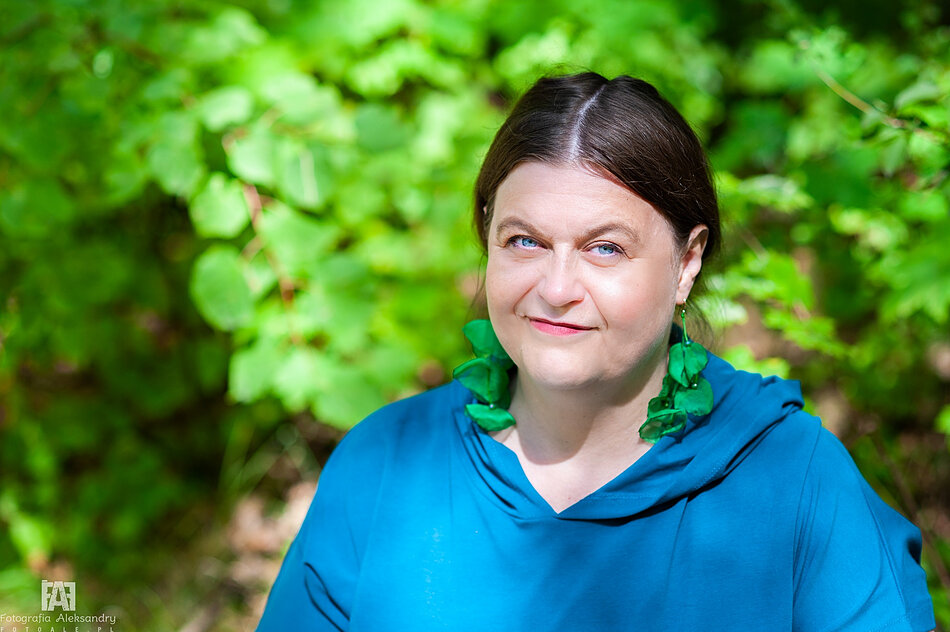 Portret osoby siedzącej na tle gęstej, zielonej roślinności. Osoba ma ciemne włosy zaczesane z przedziałkiem na środku i ubrana jest w luźną bluzę z kapturem w intensywnym niebiesko-turkusowym kolorze. Na uszach widoczne są duże, zielone kolczyki przypominające liście lub płatki roślin. Zdjęcie wykonano na zewnątrz w słoneczny dzień. Twarz i górna część sylwetki są ostre i dobrze oświetlone, natomiast tło pozostaje rozmyte, wypełnione różnymi odcieniami zieleni. W lewym dolnym rogu znajduje się znak wodny z napisem „Fotografia Aleksandry”.