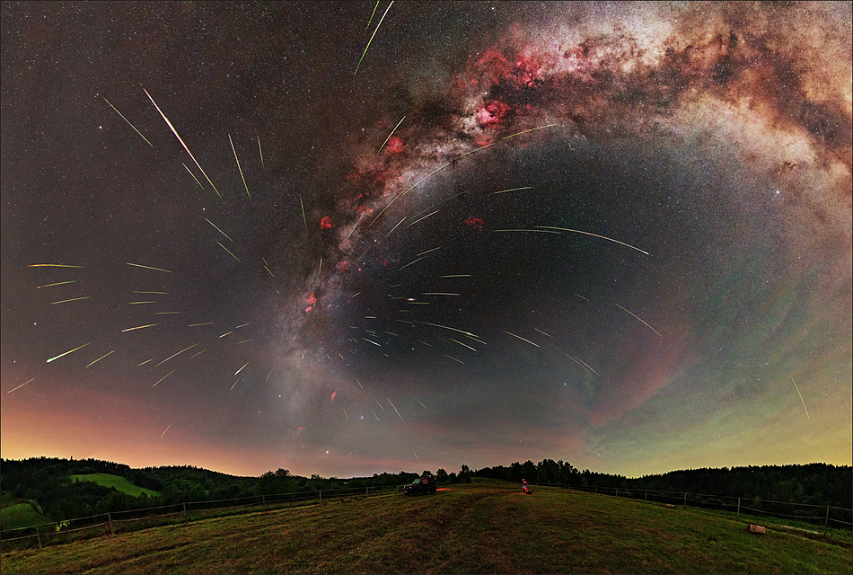 Zdjęcie przedstawia spektakularny deszcz meteorów z roju Perseidów, których ślady wydają się wybiegać z jednego punktu na nocnym niebie. W tle widoczny jest barwny, panoramiczny łuk Drogi Mlecznej rozciągający się nad ciemnym, pofałdowanym krajobrazem.