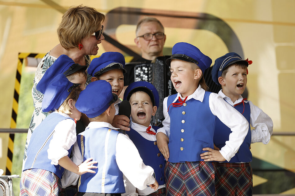 Grupa młodych chłopców i kobieta stojąca na scenie, wszyscy mają na sobie niebieskie szkolne mundurki z czerwono-białymi spódnicami i niebieskimi kapeluszami, chłopiec w środku trzyma mikrofon i wydaje się śpiewać, za nimi jest mężczyzna grający na akordeonie, a obok niego inny mężczyznę, wydaje mi się, że grupa wykonuje piosenkę lub przedstawienie.