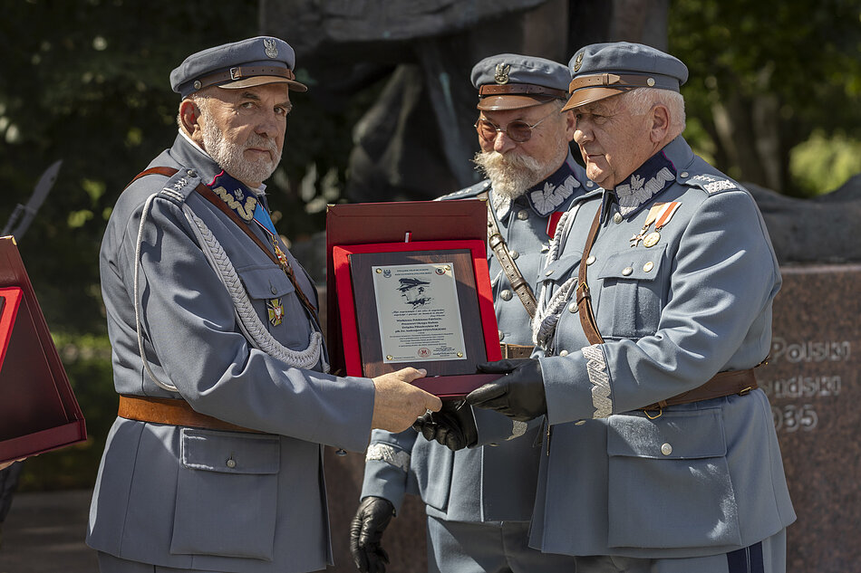 Trzy osoby w mundurach a la Piłsudski stoją razem, trzymając plakietę z wizerunkiem Marszałka