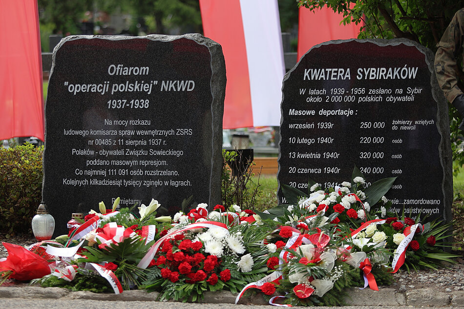Dwie czarne granitowe tablice z czerwonymi i białymi kwiatami i wstążkami umieszczonymi na nich przed czerwonym banerem. Tabliczka po lewej stronie brzmi Ofiarom operacji polskiej (NKWD) 1937-1938 a na tablicy pośrodku znajdują się nazwy KWATERA SYBIRAKOW napisane w języku polskim. Po prawej stronie znajduje się lista nazwisk i dat jest również mała biała latarnia ułożona obok cmentarza. W tle możemy zobaczyć drzewo i budynek sugerujący, że obraz został zrobiony na cmentarzu lub pomniku.