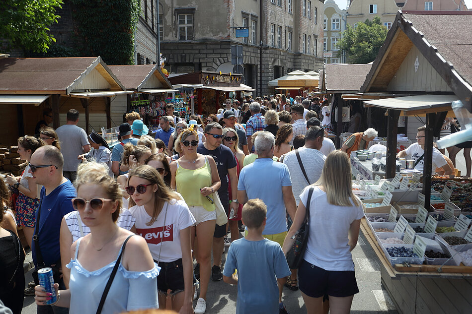 Zabiegany rynek uliczny w europejskim mieście. Rynek jest pełen ludzi w różnym wieku i pochodzenia etnicznego, którzy chodzą i przeglądają stragany po prawej stronie ulicy. Istnieje kilka straganów sprzedających różne rodzaje owoców i warzyw, w tym jabłka, pomarańcze, banany i winogrona. Niektórzy ludzie noszą okulary przeciwsłoneczne i noszą torby, podczas gdy inni noszą torebki handlowe. W tle znajdują się budynki z tradycyjną europejską architekturą i wyłożoną drzewami ulicą ze sklepami i straganami. Ludzie chodzą po obu stronach tej ulicy, ciesząc się rynkiem i atmosferą.