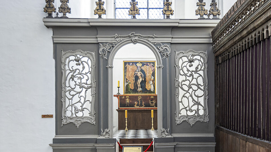 wnętrze kościoła z dużym łukiem w środku; łuk jest wykonany z szarego kamienia i ma skomplikowane rzeźby na górze i po bokach; po obu stronach ozdobione jest złotokolorowymi posągami aniołów; w tle możemy zobaczyć obraz Jezusa Chrystusa na drewnianym ołtarzu z dwoma świecami i zasłonioną na nim czerwoną tkaniną
